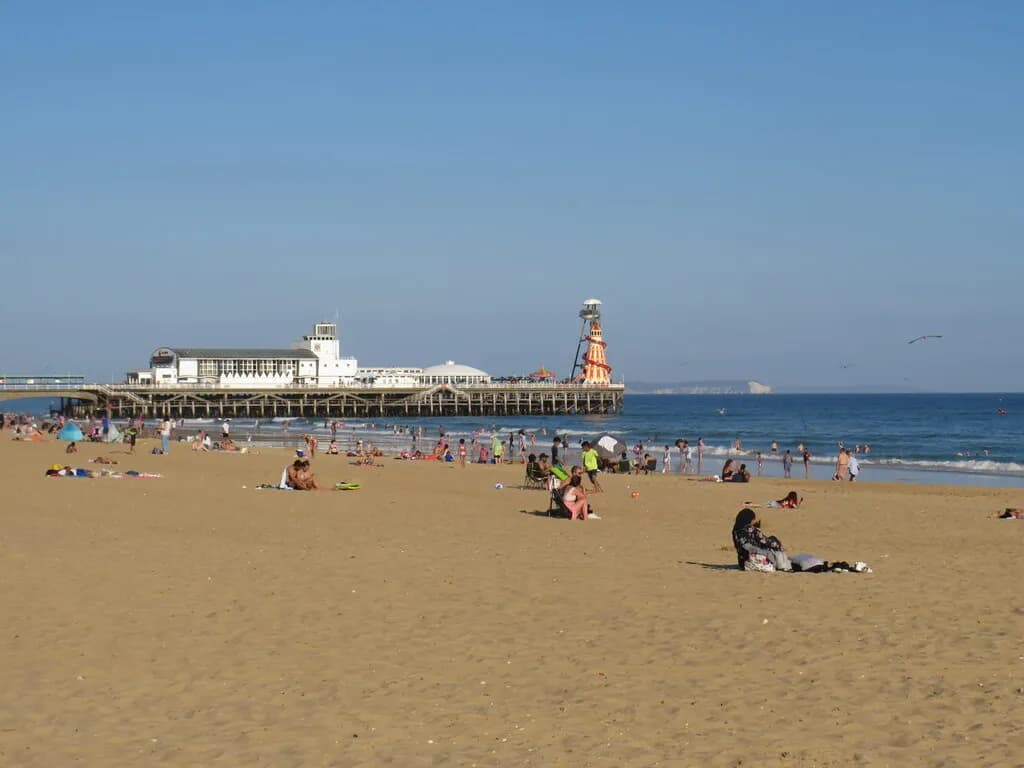 People relax and swim on a sunny sandy beach with a grand pier extending into the blue ocean in the background.